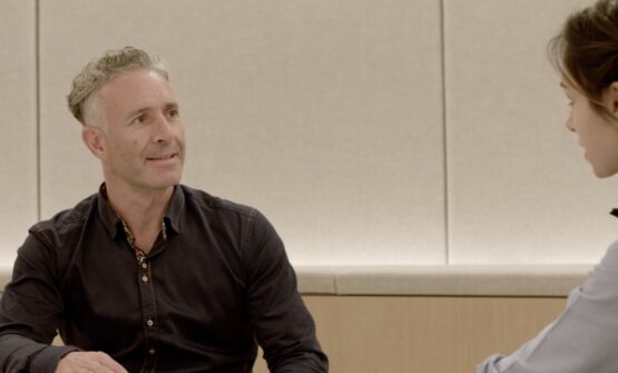 A man (Mark Nesbitt) in a meeting room, smiling and giving business advice to interested female leader